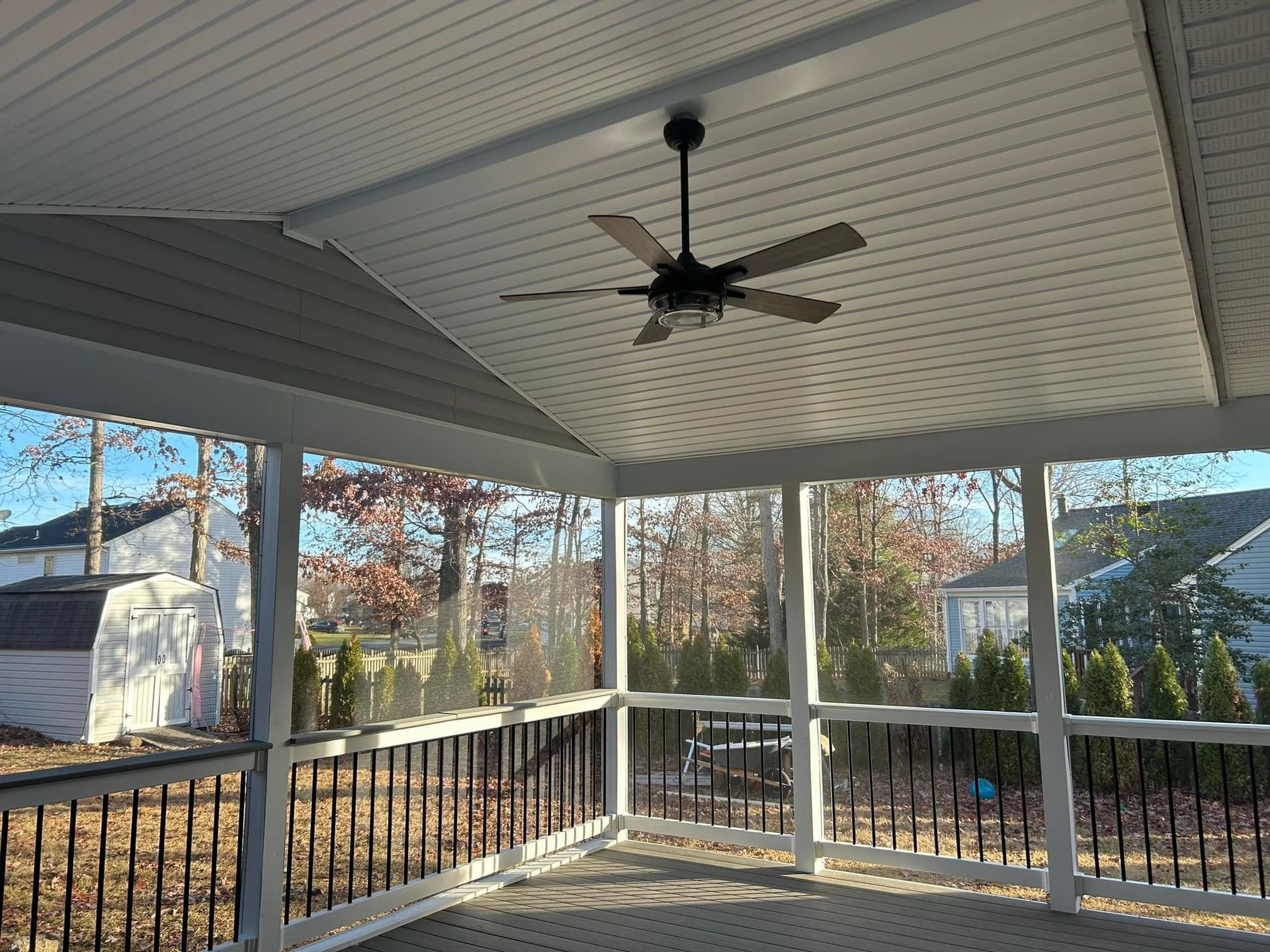 Covered porch with white ceiling, black ceiling fan, and railing overlooking trees in daylight