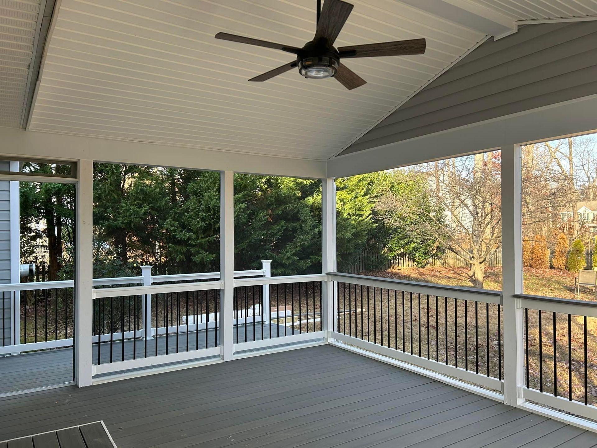 Covered porch with white railings, gray floor, and a ceiling fan overlooking trees in autumn.