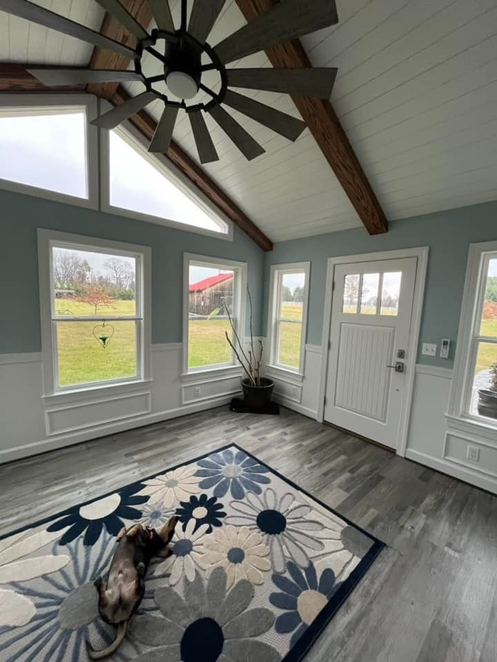Bright sunroom with vaulted ceiling, large windows, blue floral rug, and a small dog lying on the floor.