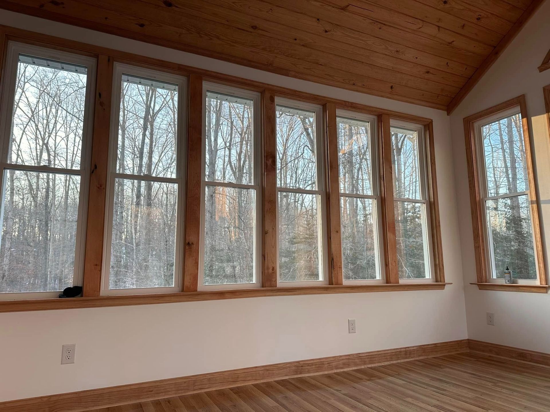 Empty sunroom with large windows, wood ceiling, and hardwood floor overlooking leafless trees.