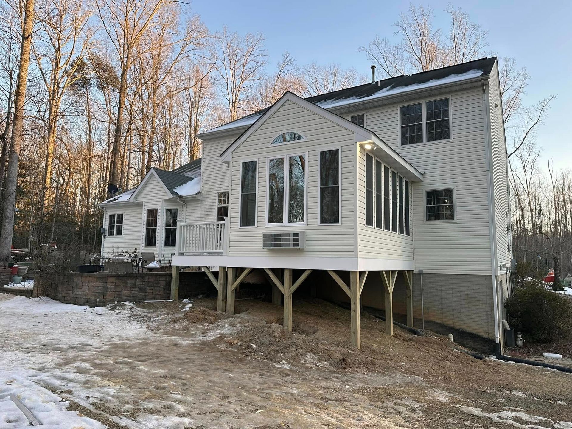 Snowy suburban house with white siding and black shutters, raised on stilts in a wooded yard