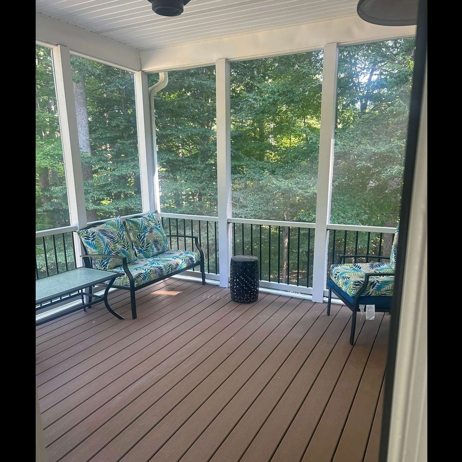 Screened porch with brown deck flooring, black metal seating, and green trees beyond the railing.