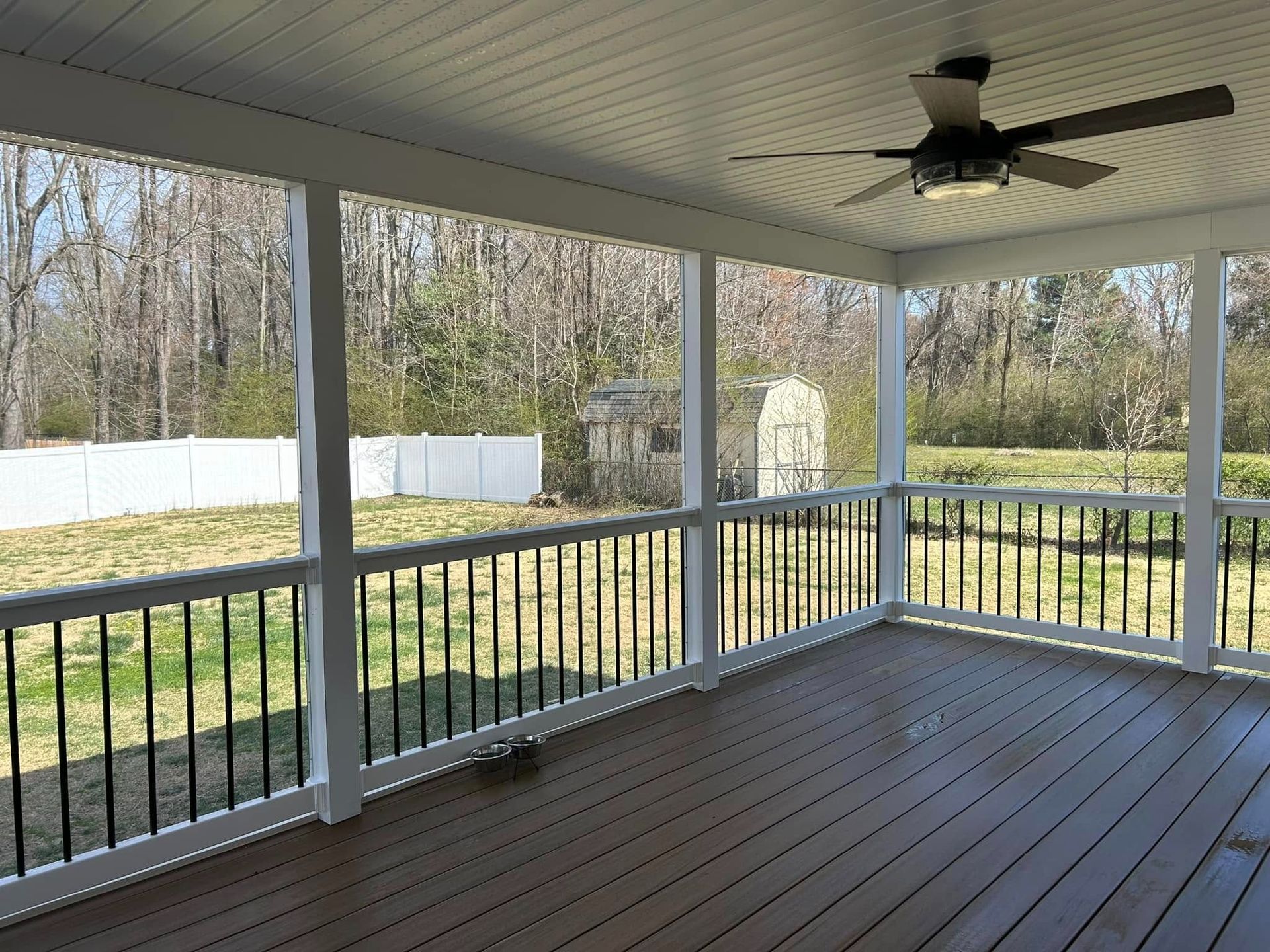 Covered porch with white railings, brown floorboards, and a ceiling fan overlooking a grassy yard and trees.