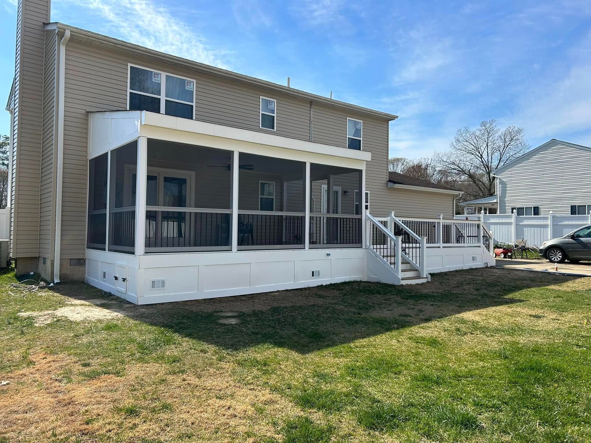 Backyard deck attached to a beige house with white railings and screened porch, under a blue sky