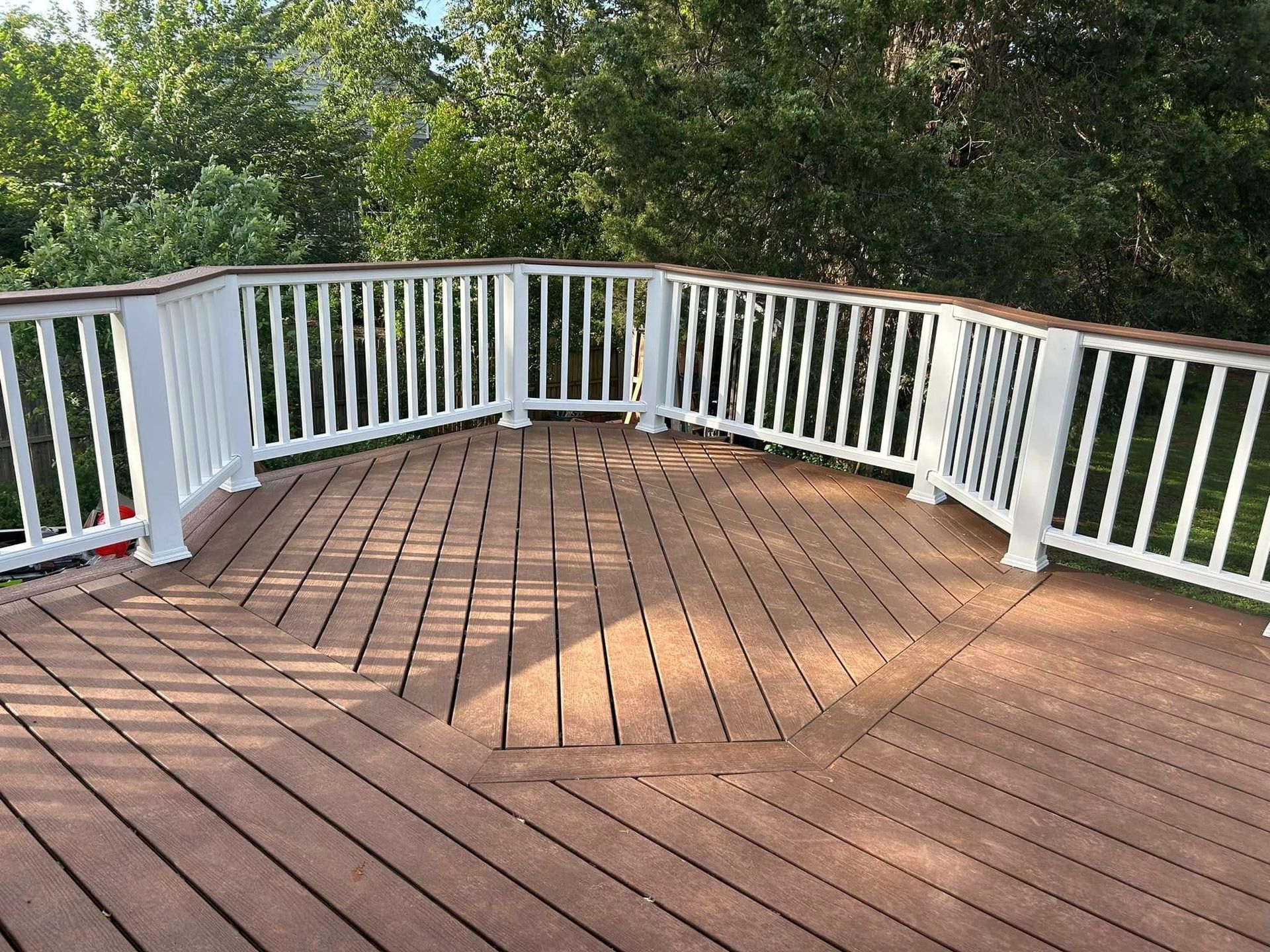 Wooden deck with white railing overlooking green trees, lit by sunlight and shadows