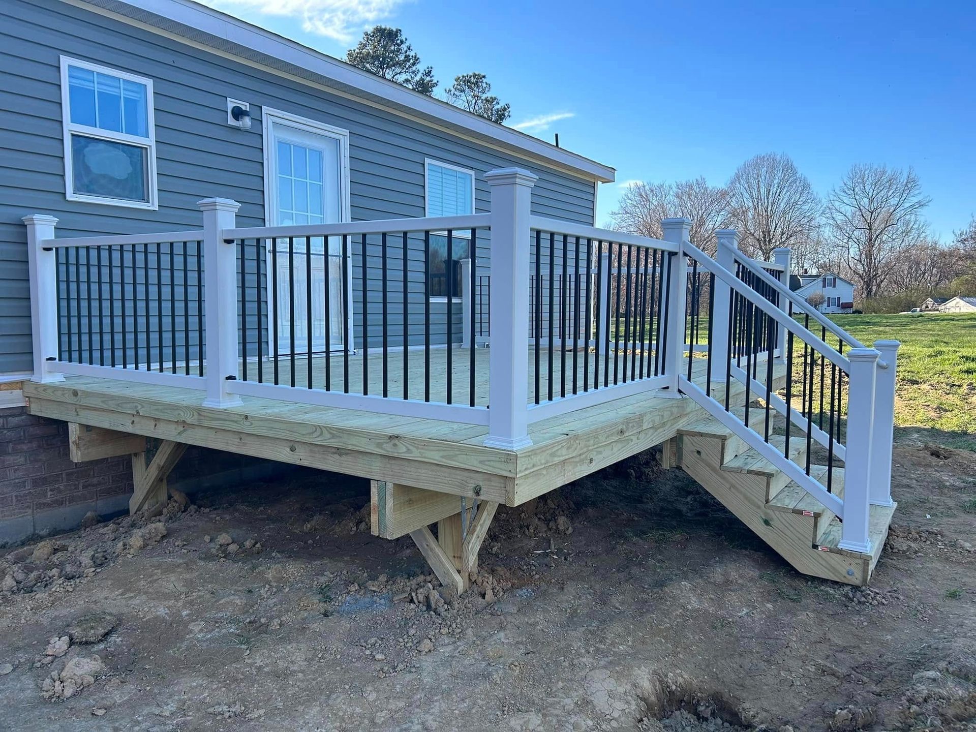 Elevated white-and-gray deck with railing and stairs attached to a house on a dirt yard
