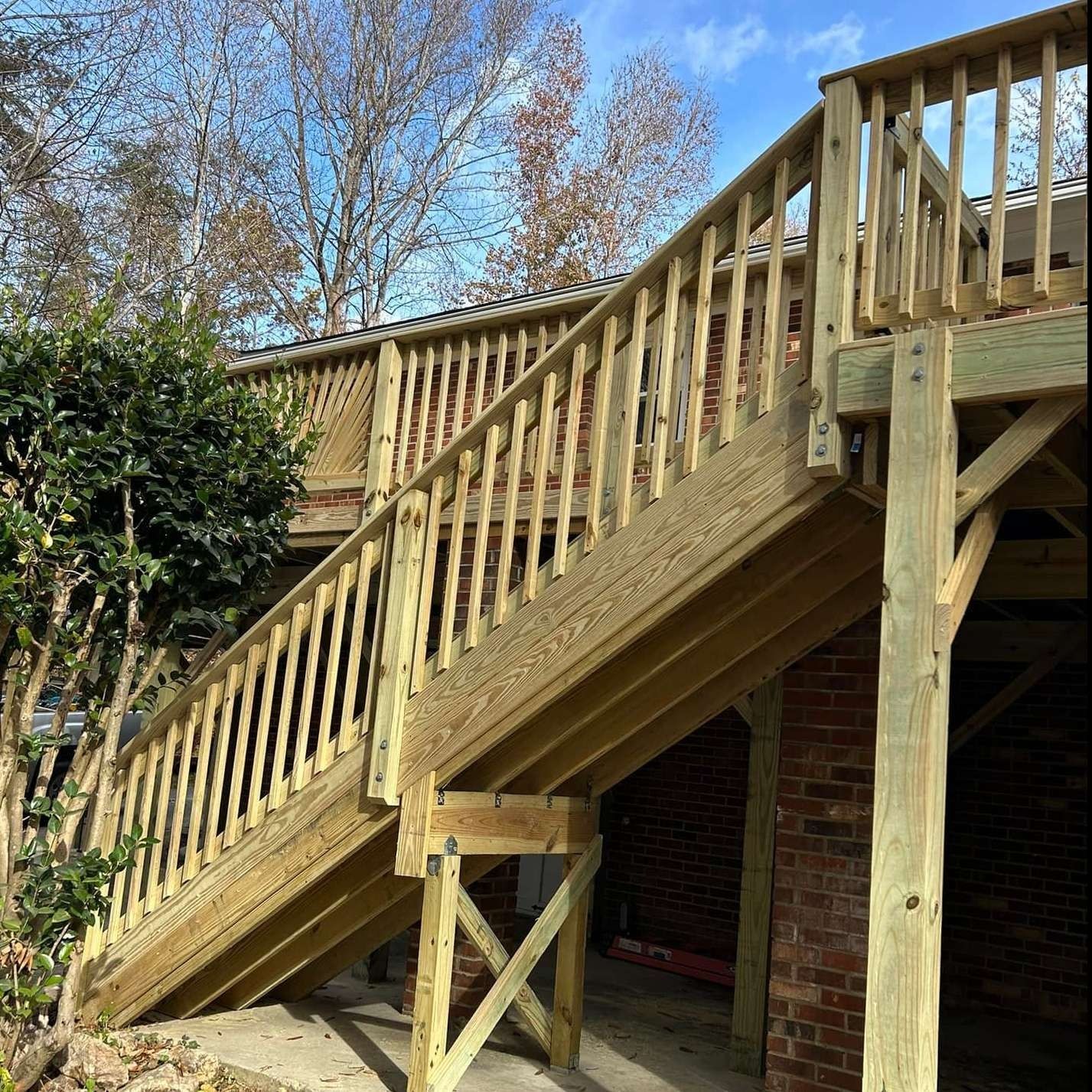 Wooden staircase and deck with railings beside a house, framed by leafless trees and a shrub.