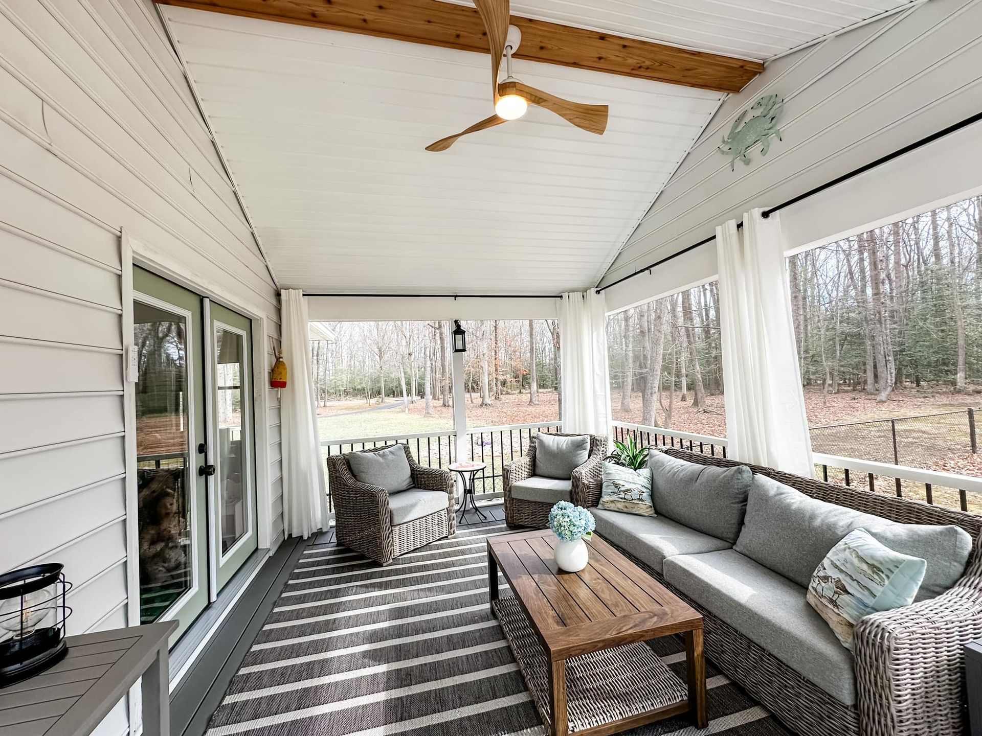 Covered porch seating area with gray sofa, wicker chairs, and a coffee table overlooking trees
