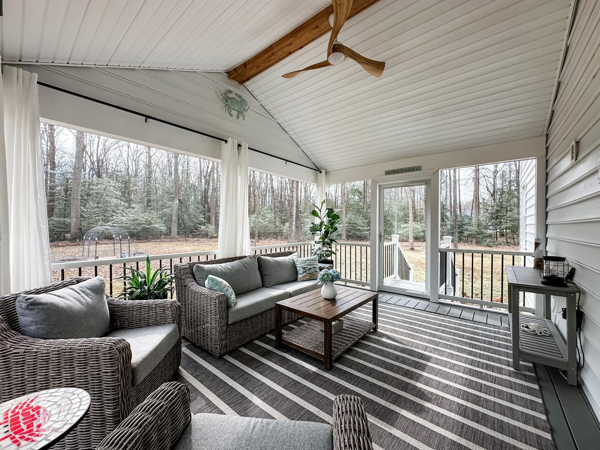 Covered porch with wicker seating, striped rug, ceiling fan, and wooded view beyond railings