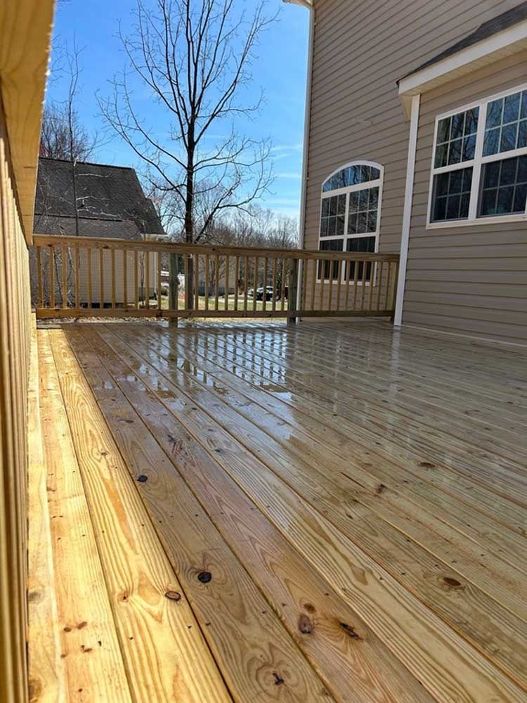 Wet wooden deck beside beige house with railing, trees, and blue sky beyond.