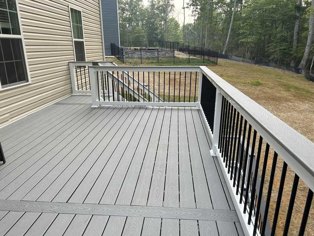 Empty gray deck with black railings beside a beige house, overlooking a wooded yard.