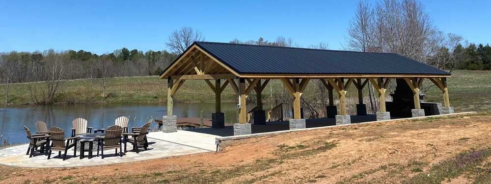 Open-sided wooden pavilion by a pond with picnic tables and chairs on a sunny day