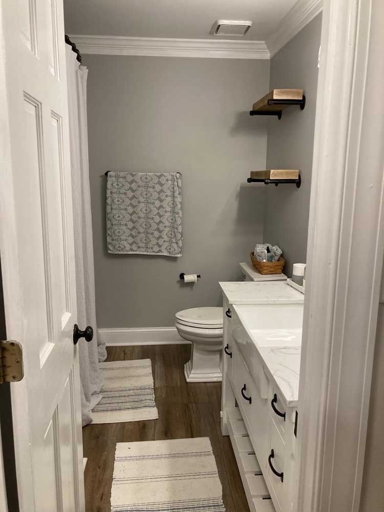 Small bathroom with white vanity, toilet, gray walls, wood floor, and floating shelves.