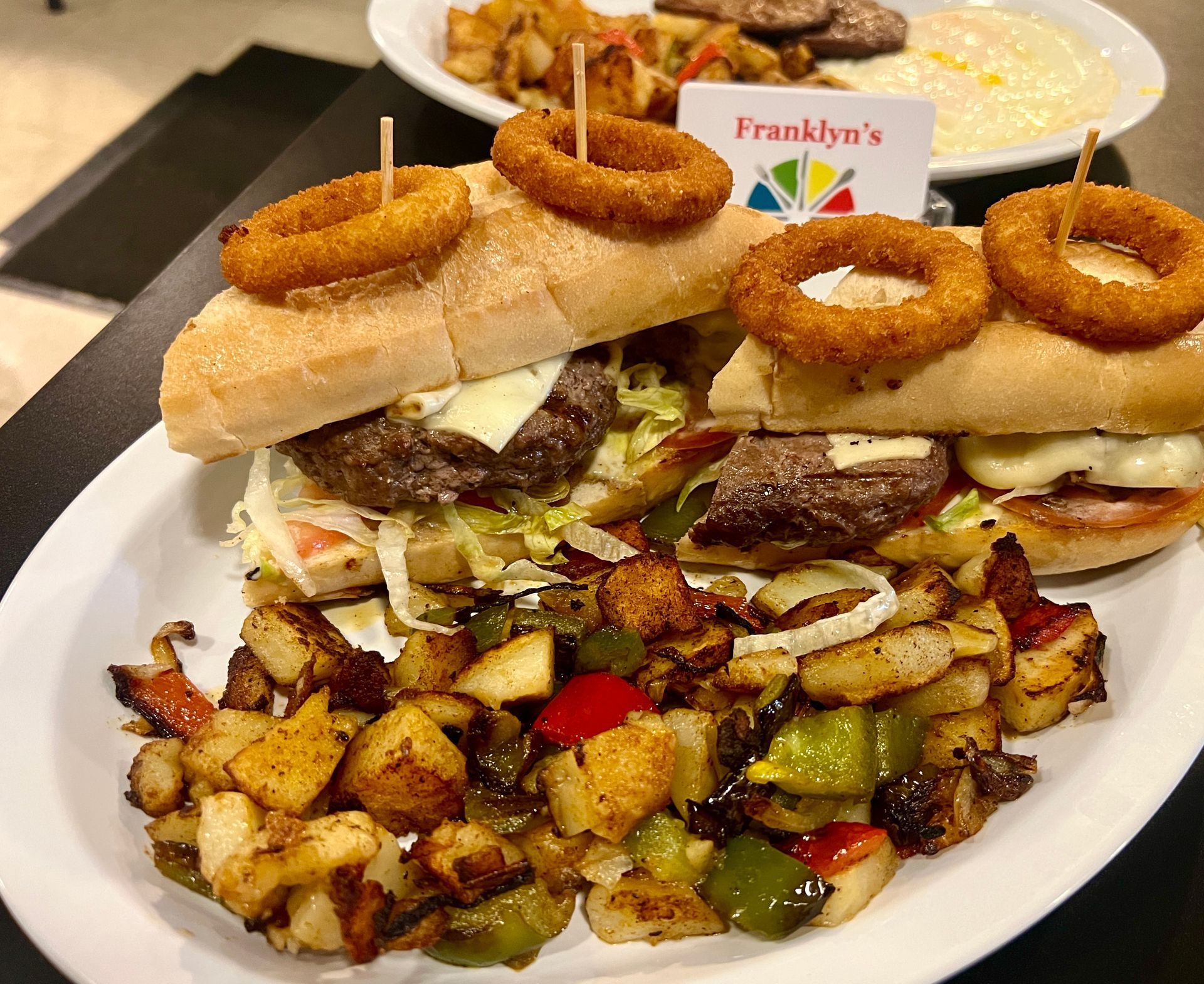 Plated sandwiches topped with onion rings and roasted vegetables, with a restaurant sign in the background
