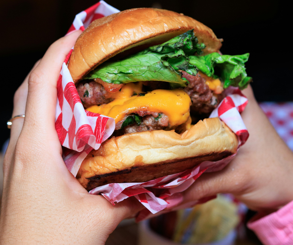 Hands holding a cheeseburger wrapped in red-and-white paper, with lettuce and melted cheese visible.