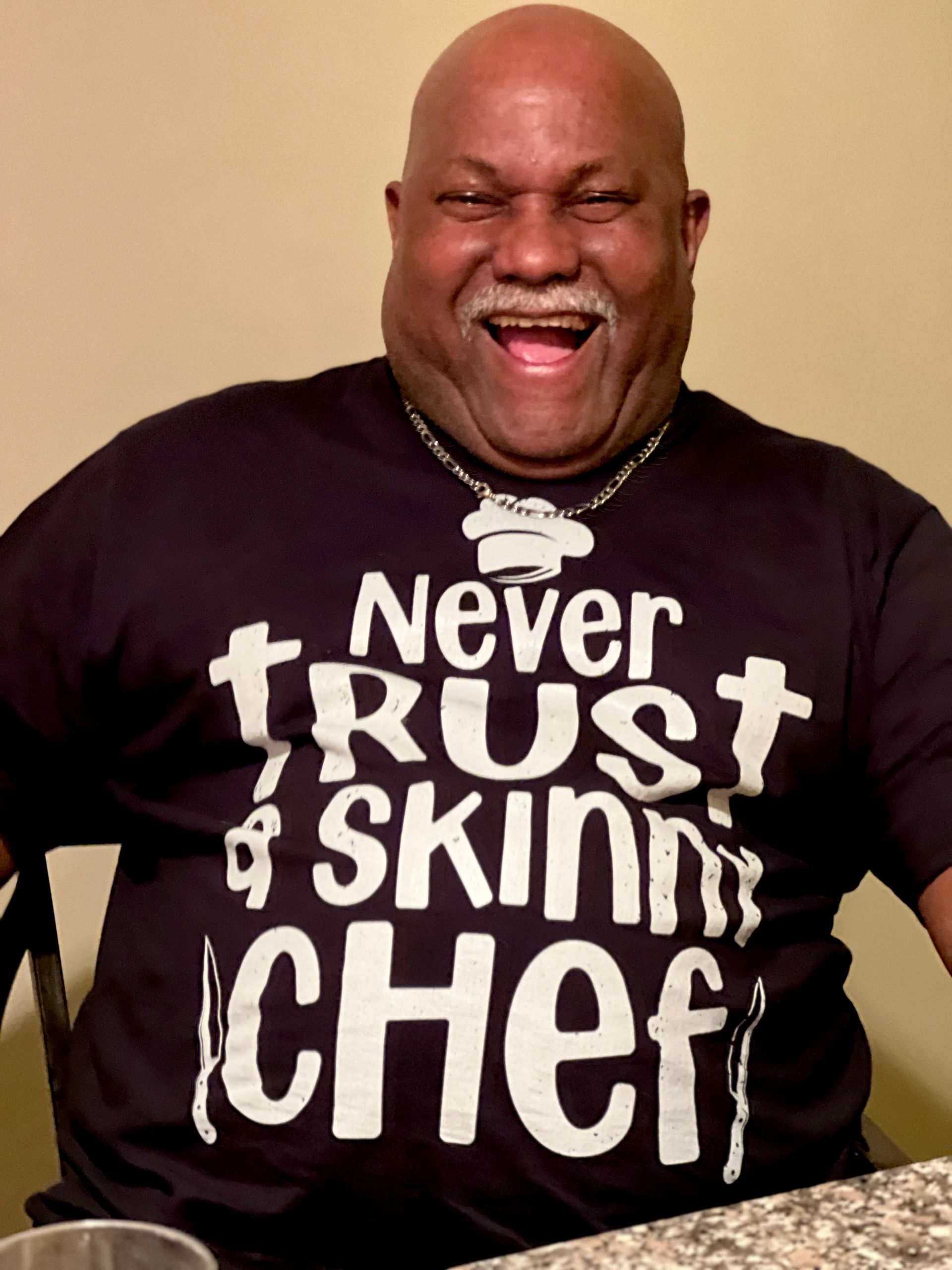 Smiling man in black shirt reading “Never trust a skinny chef” with a chef’s hat graphic