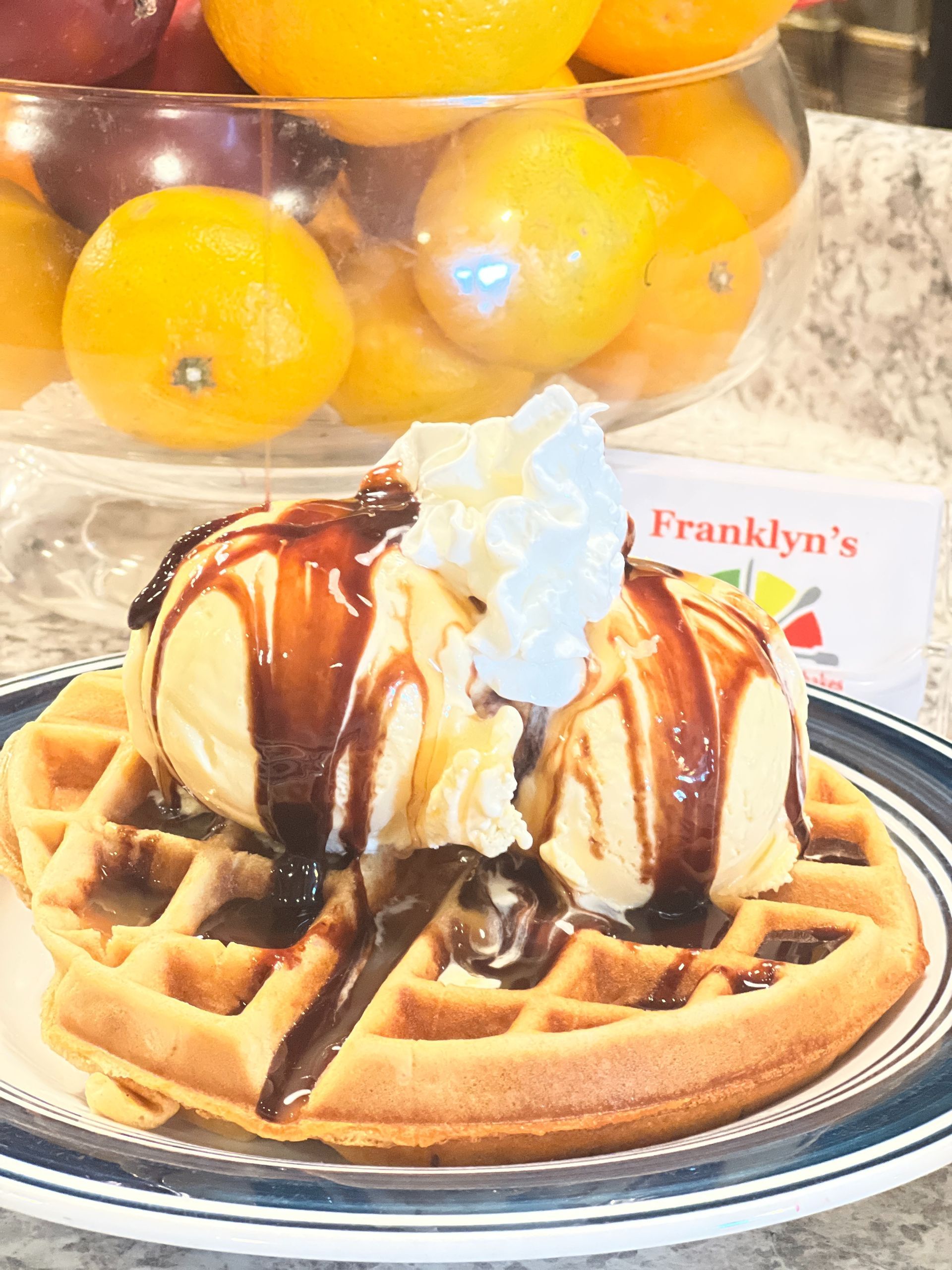 Waffle with whipped cream and chocolate drizzle on a plate, with oranges and a Franklyn’s sign behind it