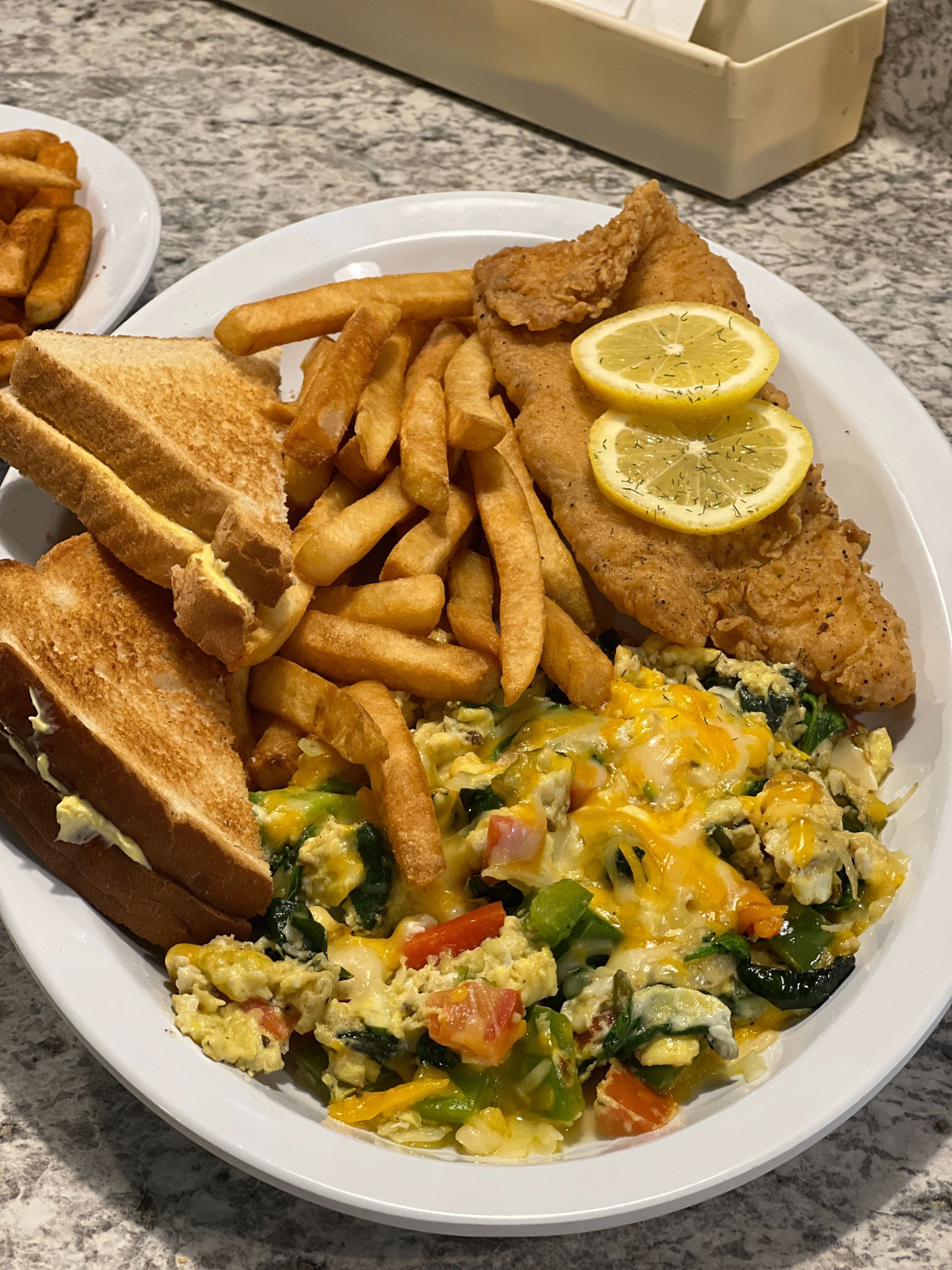 Plate with fried fish, fries, cheesy vegetables, and toast with lemon slices on a table