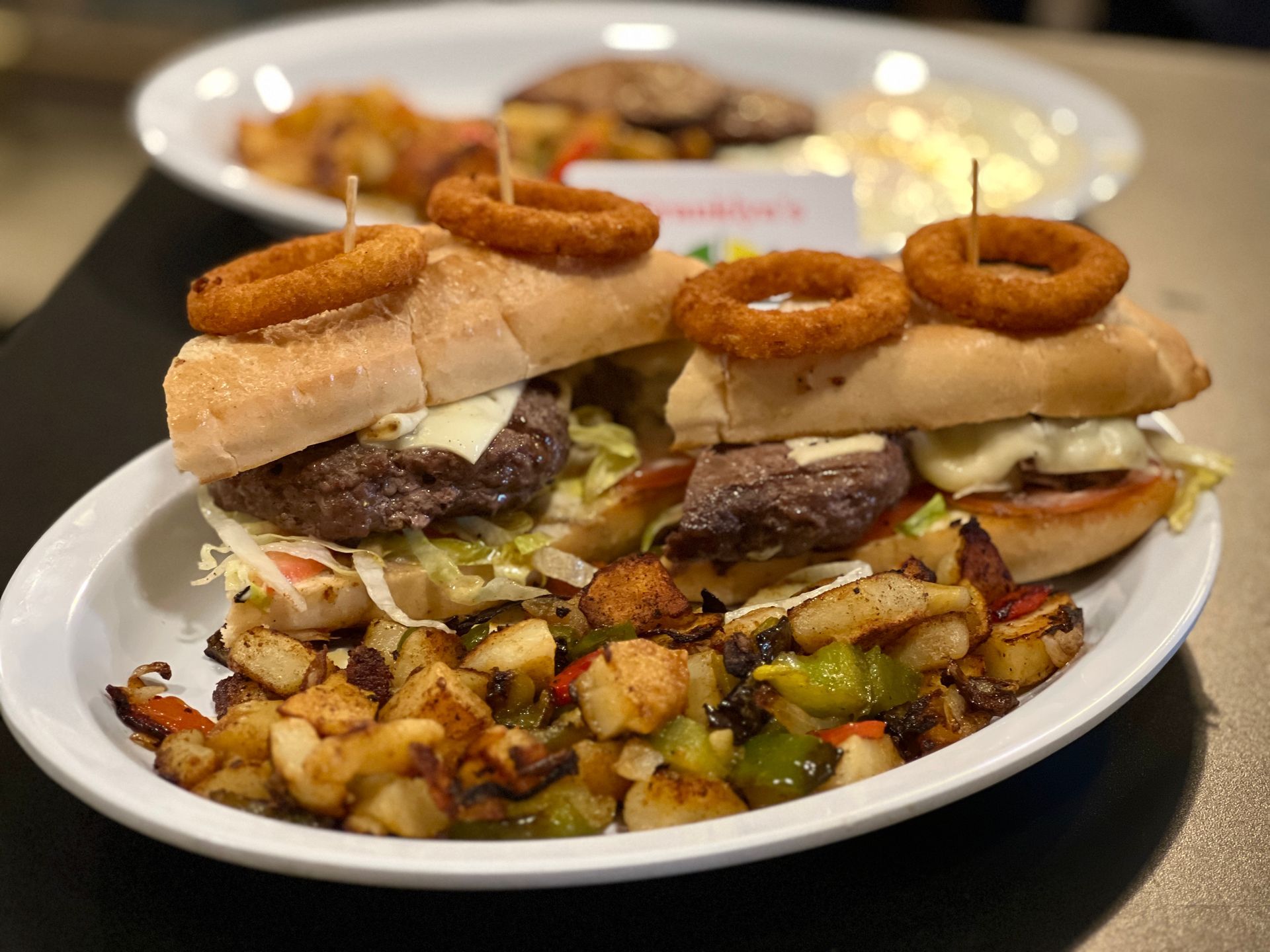 Two sandwiches topped with onion rings on white plates, with roasted potatoes in the foreground.