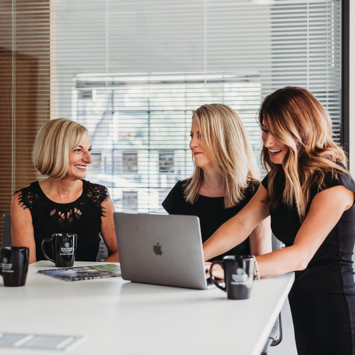 Three women are sitting at a table looking at an apple laptop