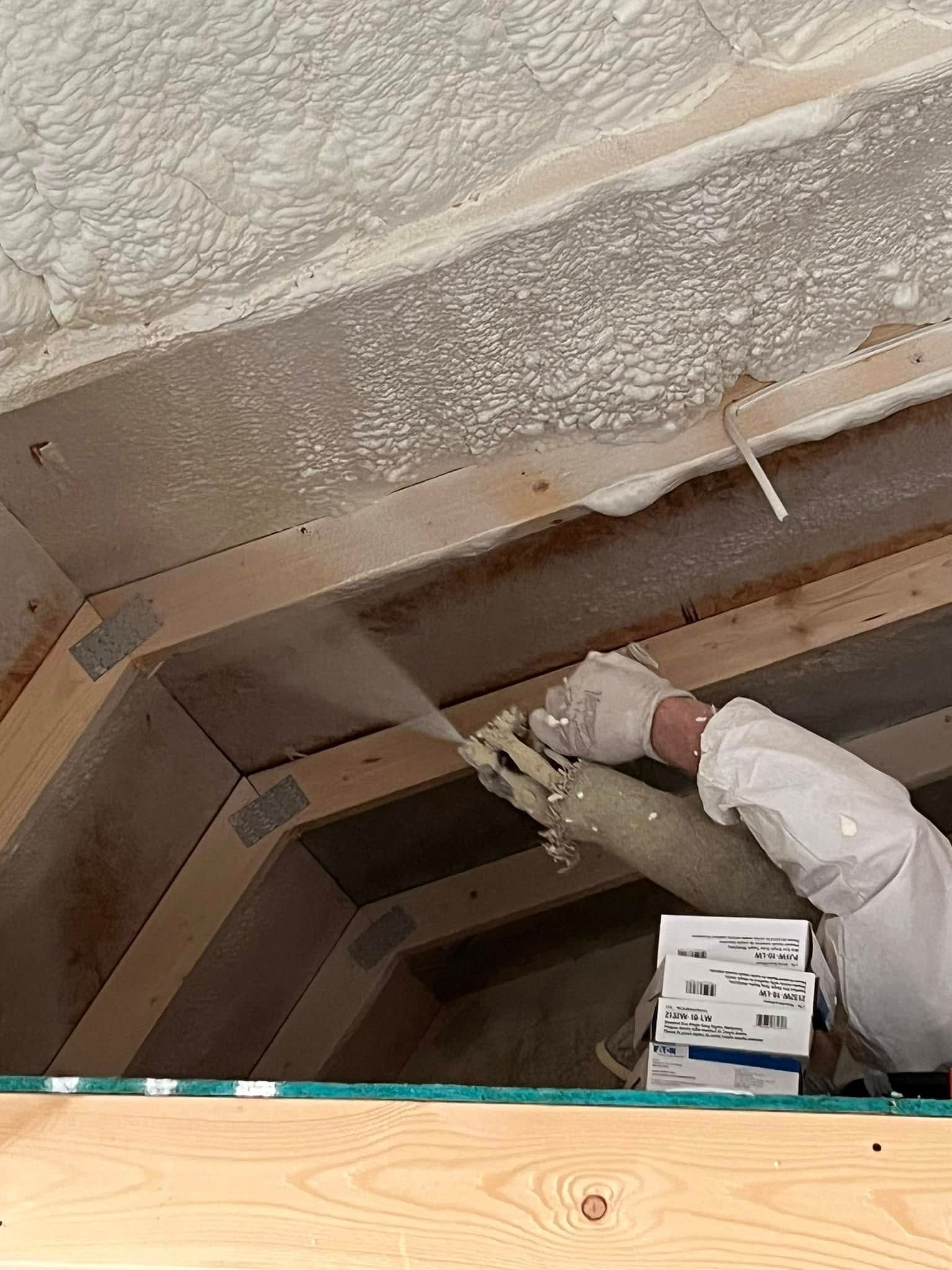 Worker spray-foaming the underside of a roof or ceiling inside a wooden-framed space.