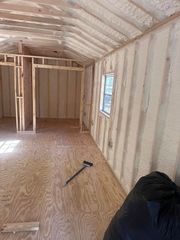 Unfinished insulated room with exposed framing, plywood floor, small window, and tools on the floor.