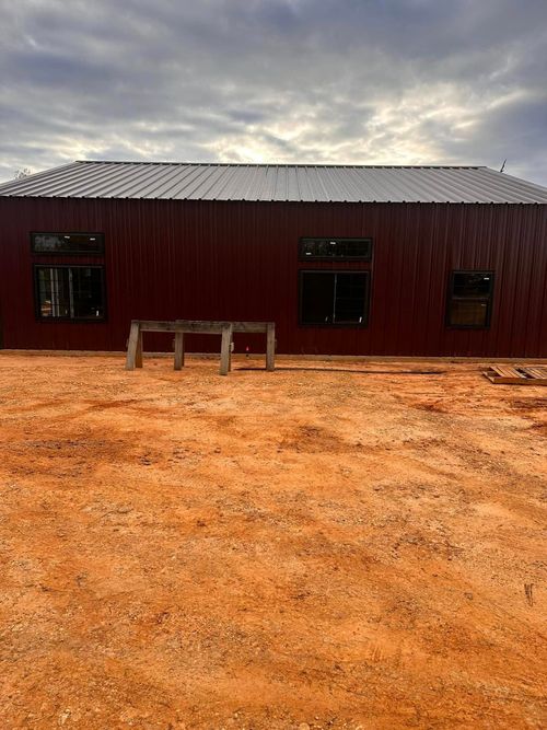 Red wooden building with a metal roof on a dirt lot under a cloudy sky.