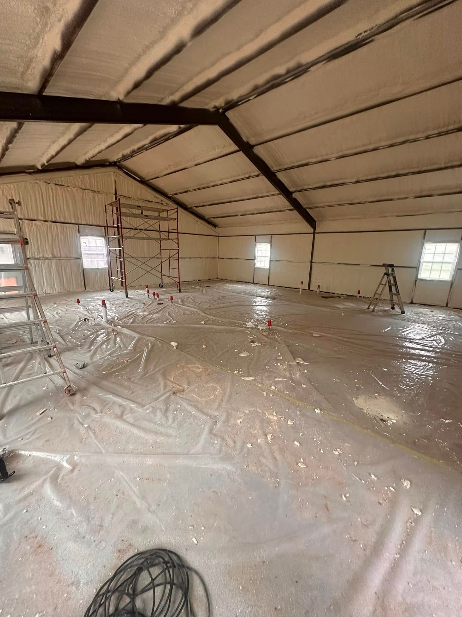 Empty unfinished attic with insulated roof, clear plastic floor covering, and exposed beams and windows.