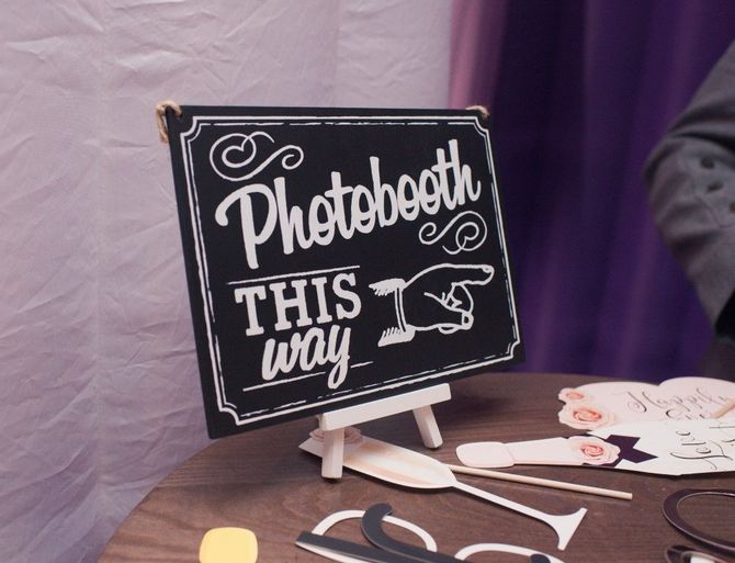 Handmade sign reads “Photobooth this way” on a table beside purple drapes and cutout props.