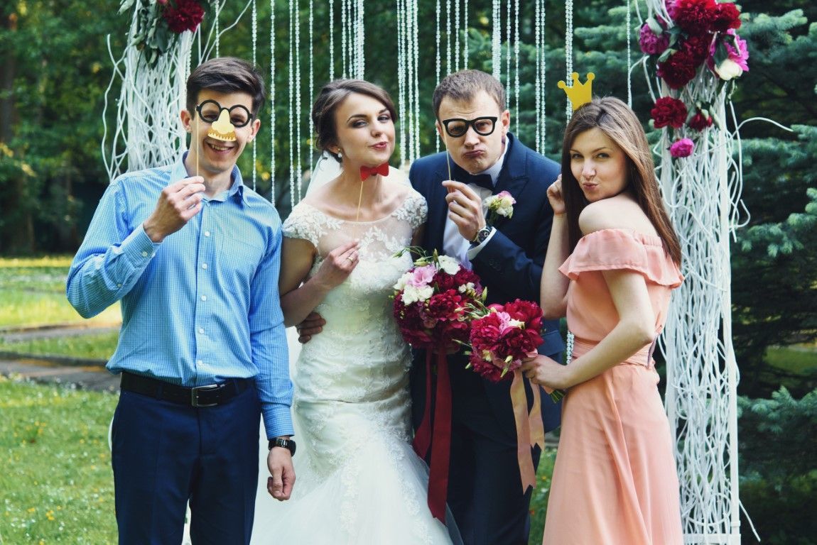 Four people posing outdoors in formal attire, holding flowers in a garden with white arch decor.
