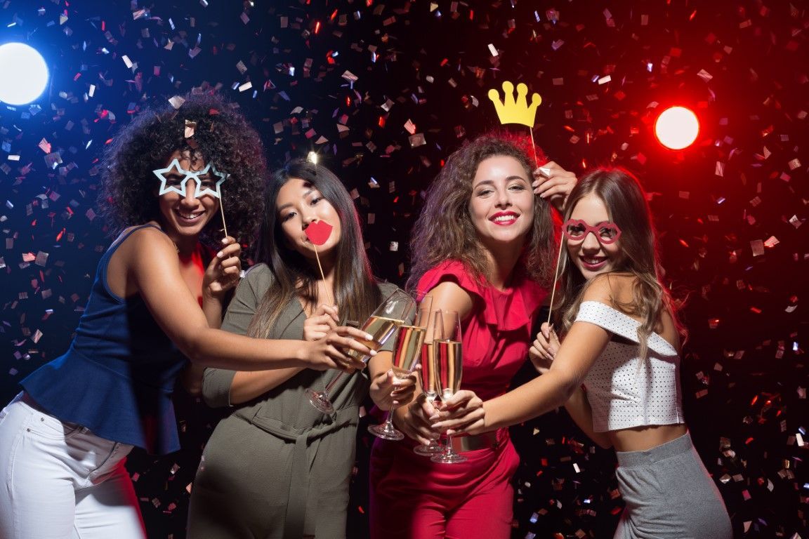 Four friends in party costumes holding champagne glasses amid confetti and colorful lights