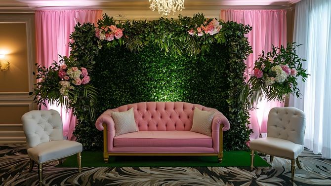 Pink tufted sofa between white chairs before a lush floral green wall with pink drapes in an elegant event setup
