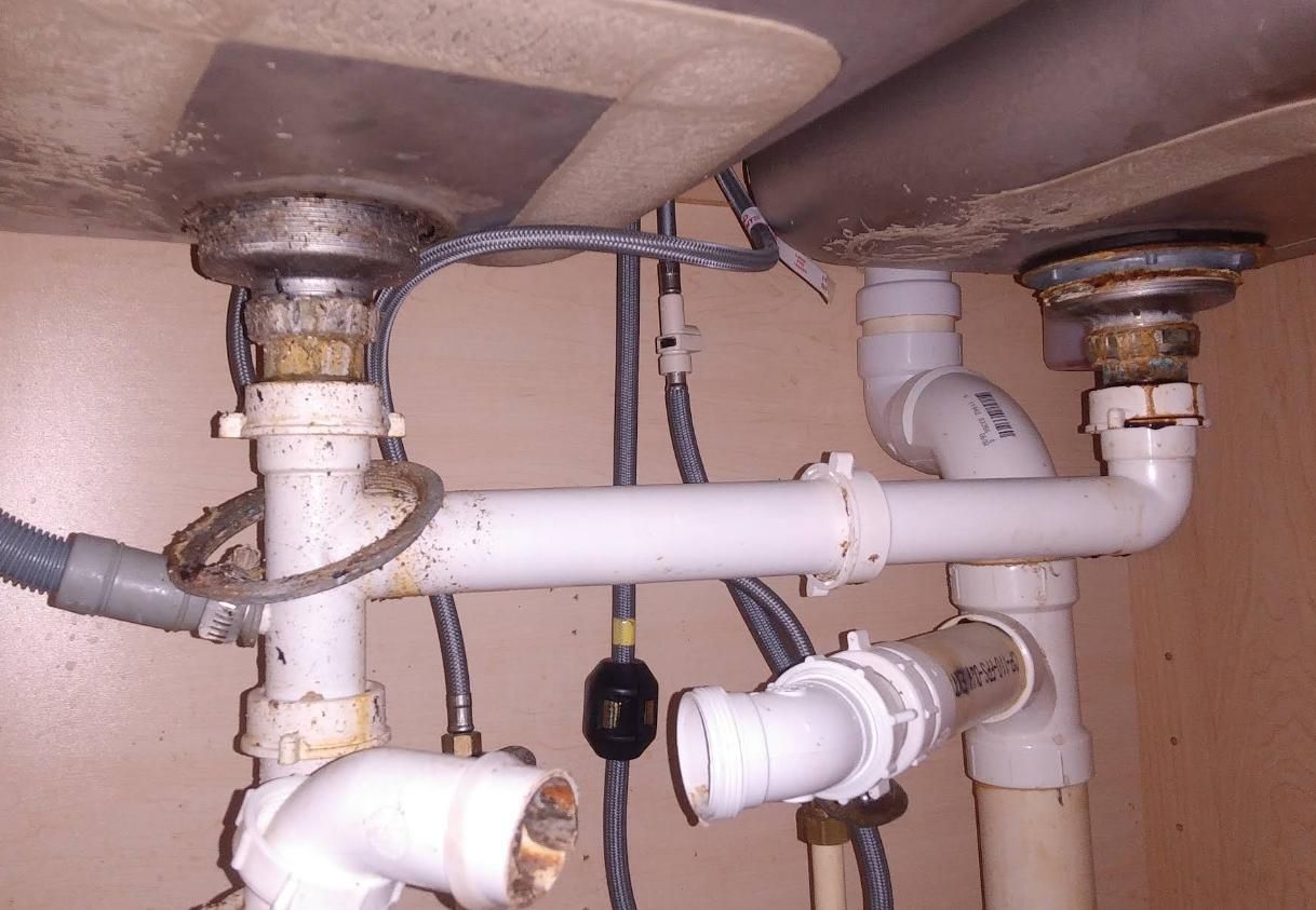 Plumbing beneath a double kitchen sink with white PVC pipes, connecting drains.