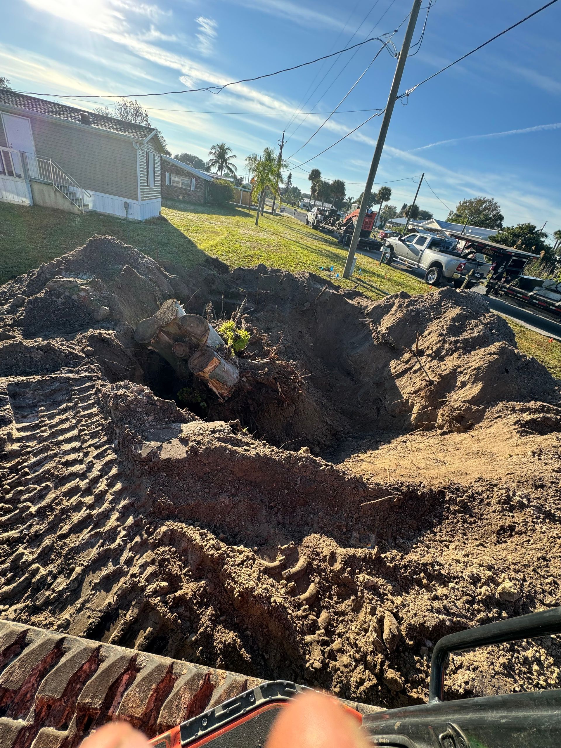 Large dirt excavation pit beside a road with a yellow excavator arm and tire tracks in the foreground