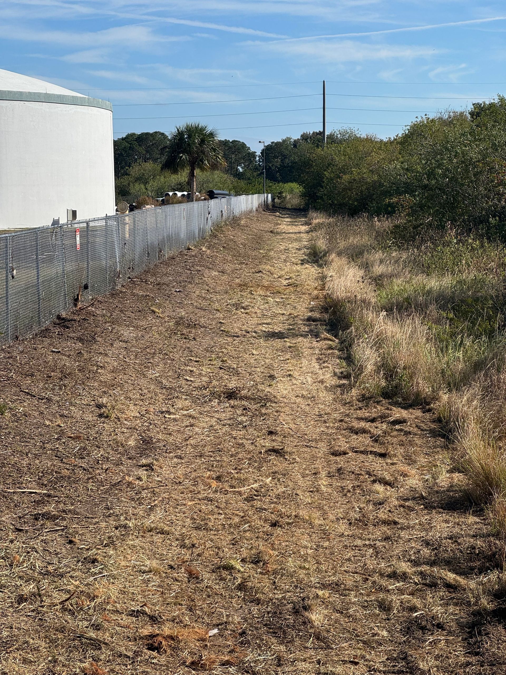 Dirt path beside a white building and chain-link fence, with dry brush and trees under a clear blue sky.
