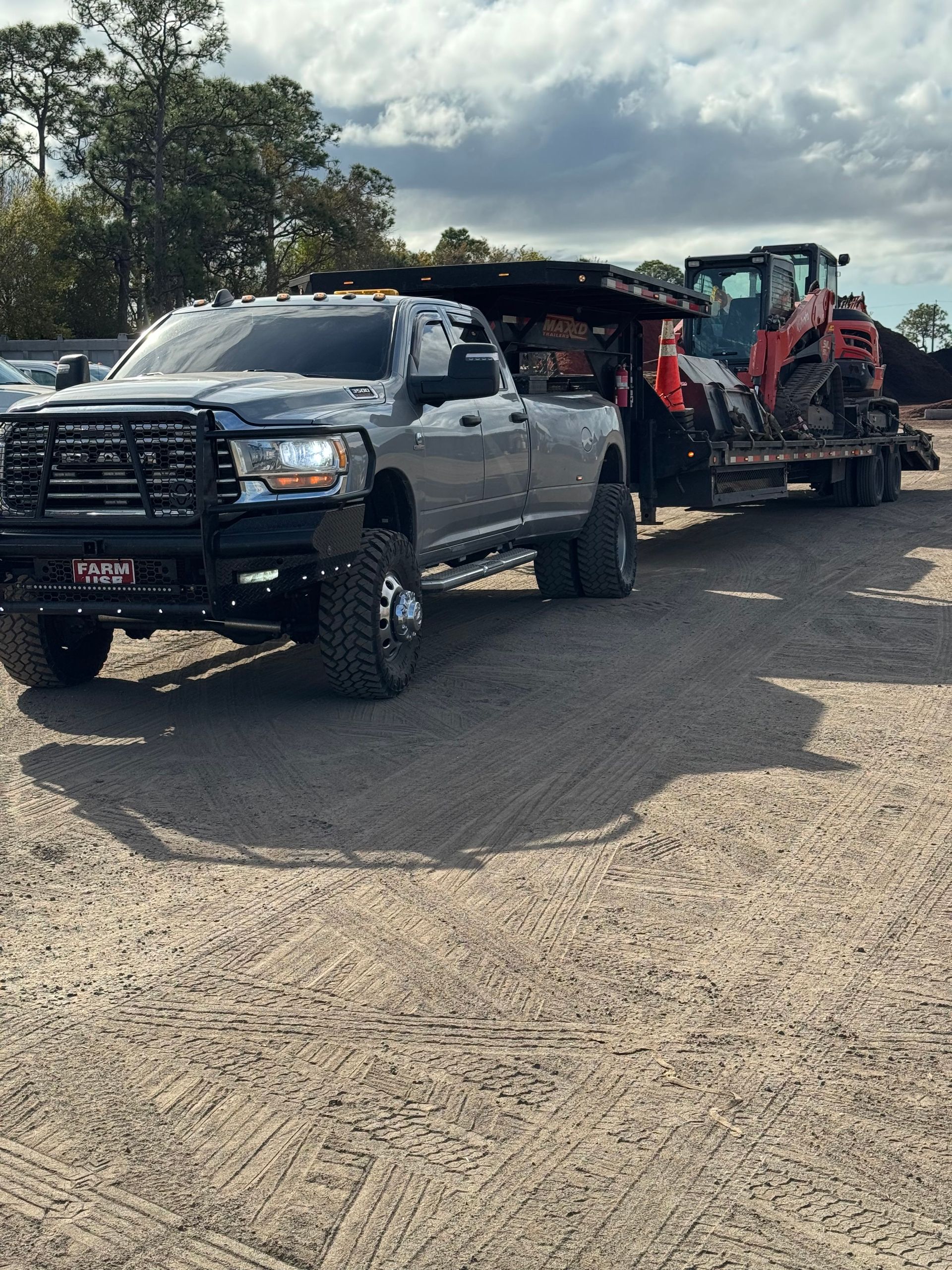 Black pickup truck towing red construction equipment on a flatbed trailer in a gravel lot