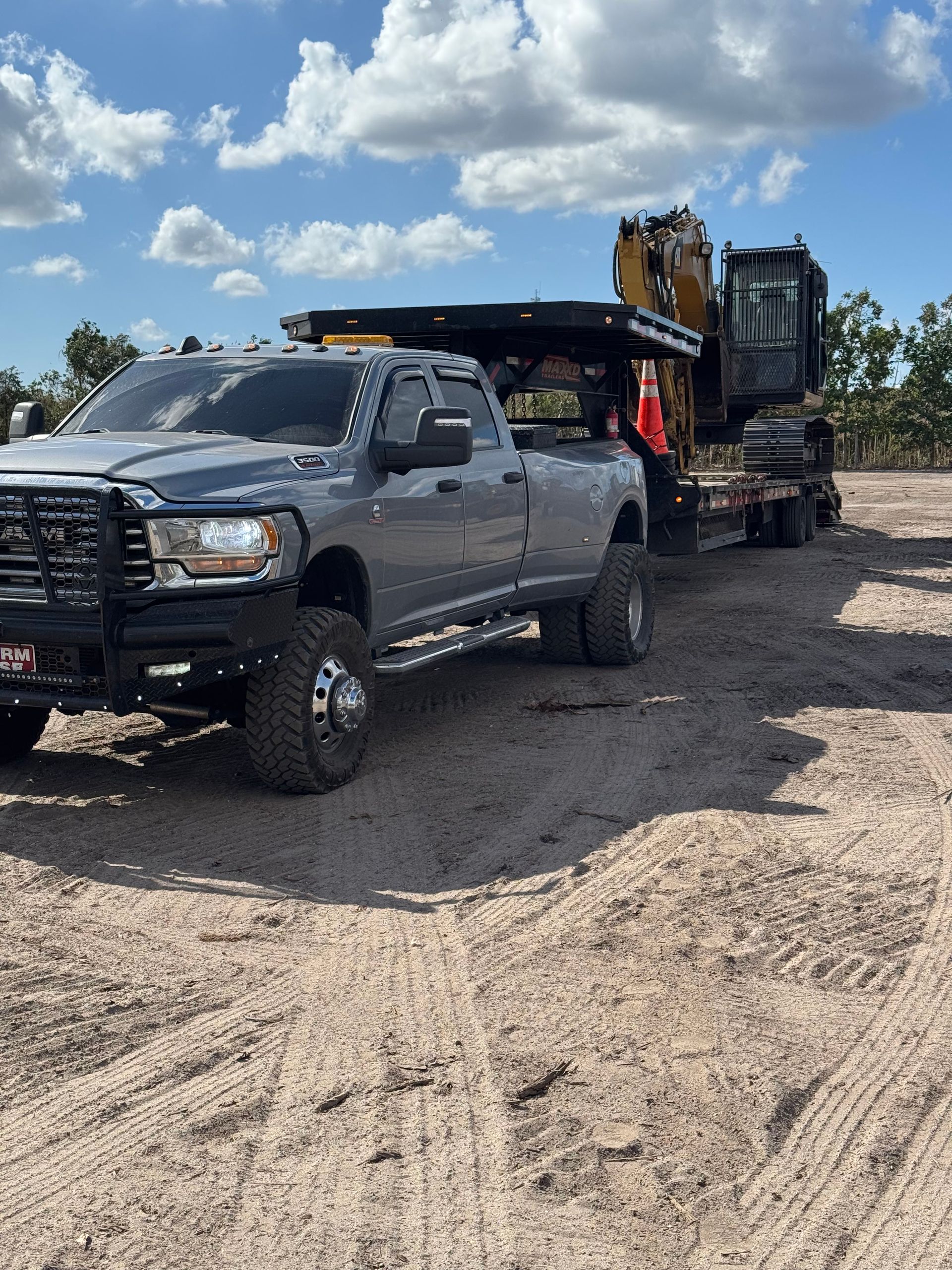 Black lifted pickup truck hauling a large excavator on a trailer in a gravel lot under a partly cloudy sky