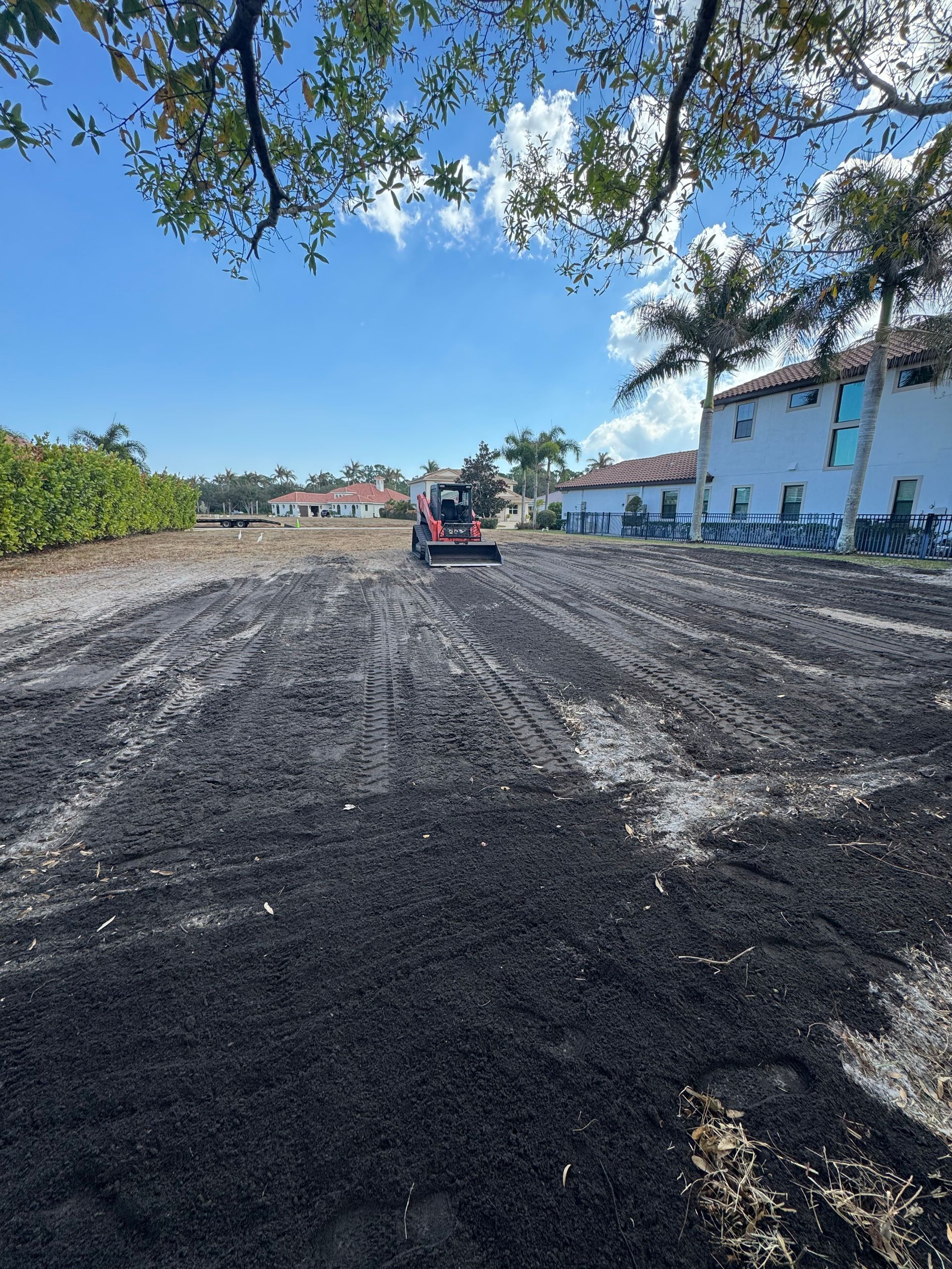 Construction vehicle grading a muddy lot beside white buildings under a blue sky