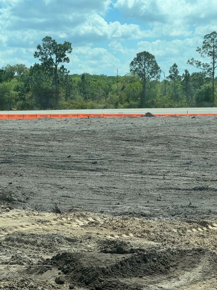 Flat dirt lot beside a road with an orange barrier, trees, and a cloudy blue sky.