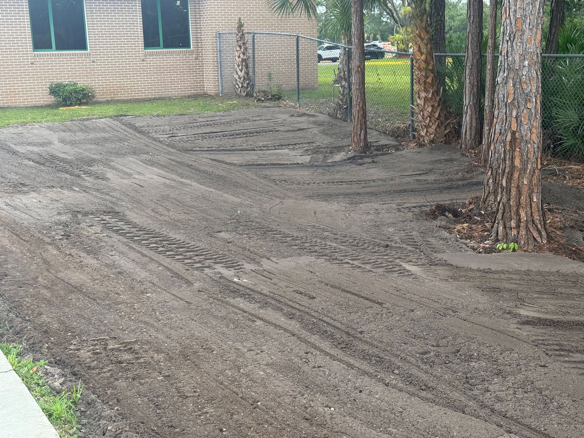 Dirt-covered yard beside a brick building and trees, with fresh tire tracks in the soil.