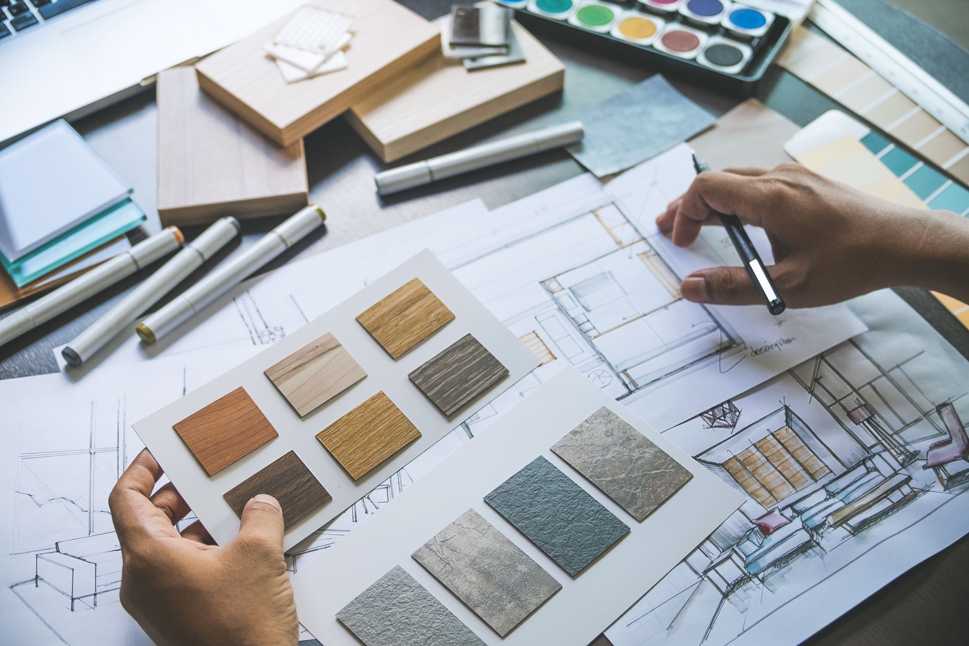 Les mains d'une personne tenant des échantillons de matériaux au-dessus de plans et d'outils de conception posés sur une table.