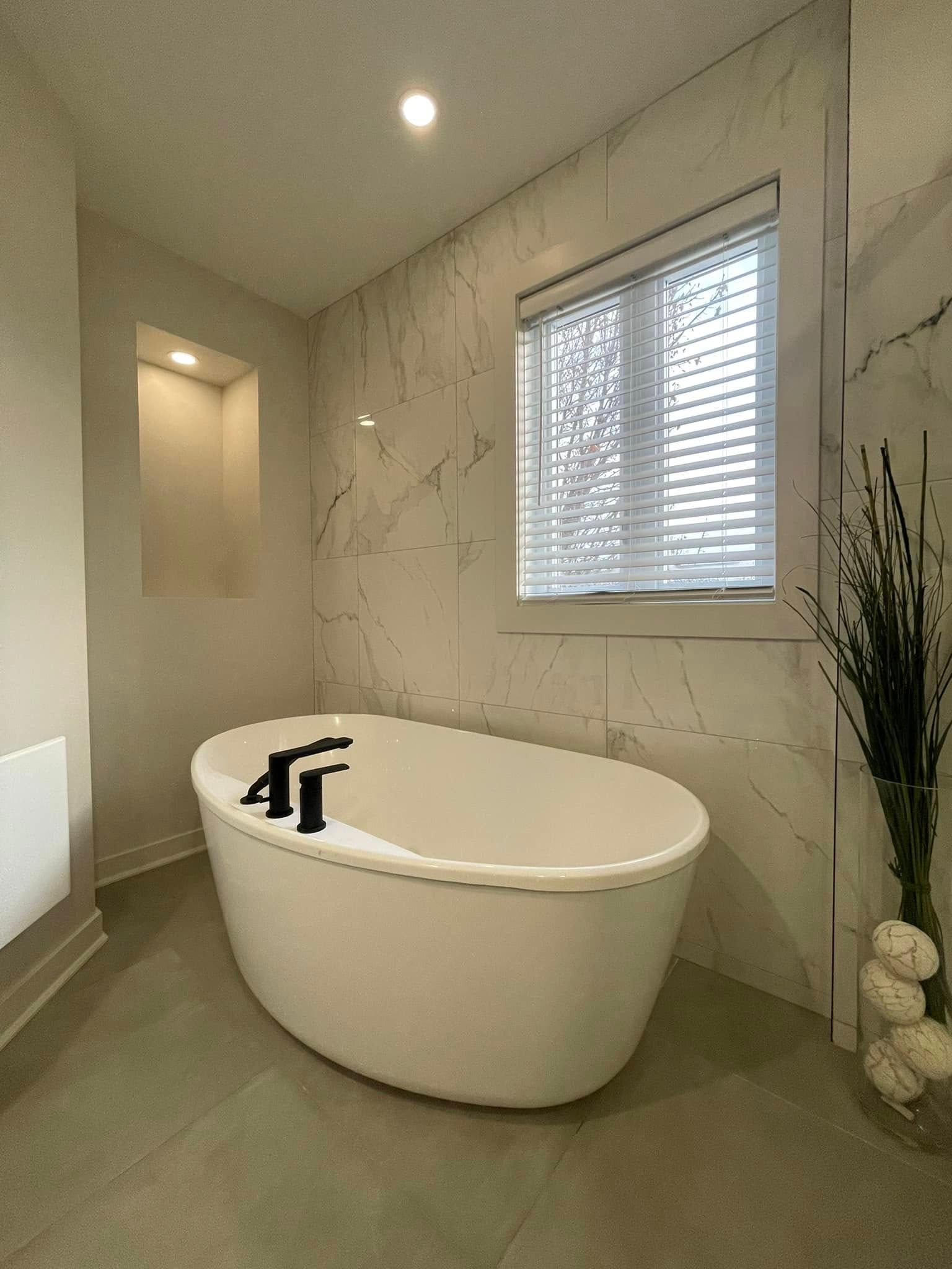 Salle de bains moderne avec baignoire îlot, carrelage en marbre, fenêtre avec stores et vase décoratif.