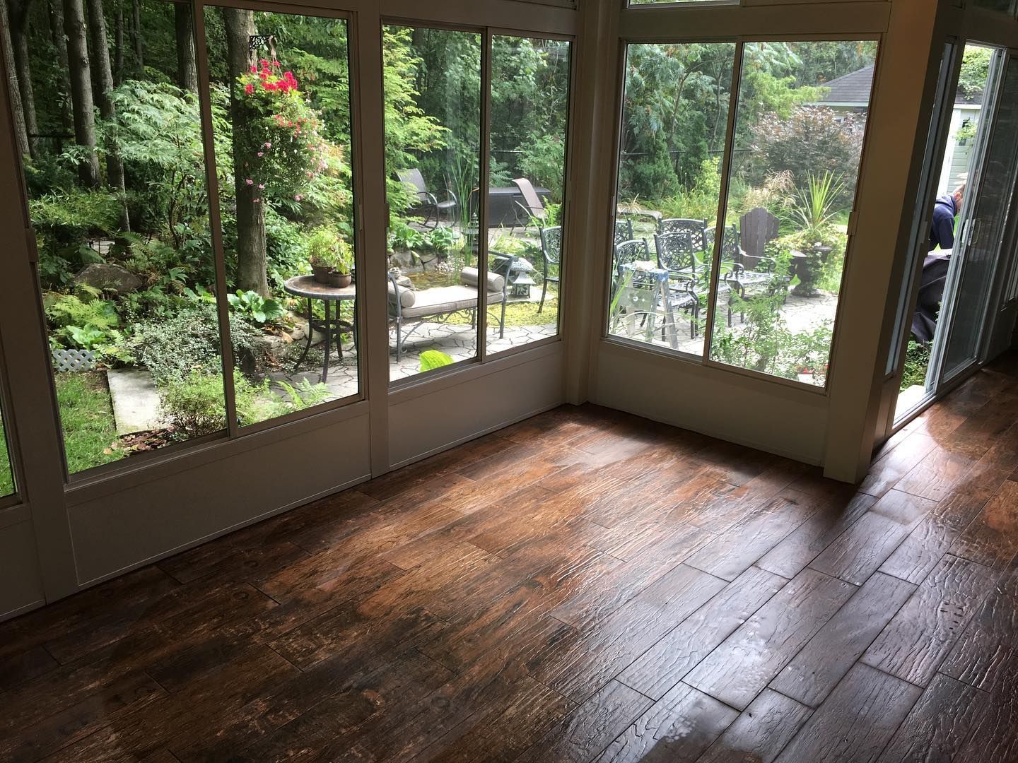 Intérieur d'une véranda avec sol en bois sombre et humide, cadres de fenêtres blancs et vue sur un jardin verdoyant luxuriant.