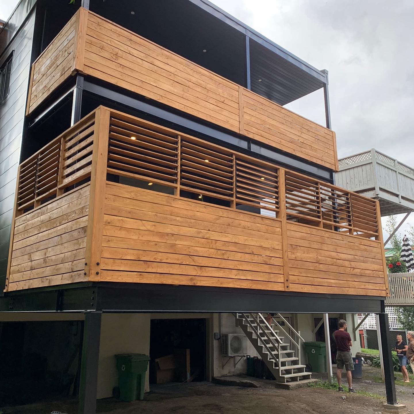Terrasse en bois à plusieurs niveaux attenante à un bâtiment, avec escaliers et personnes visibles.