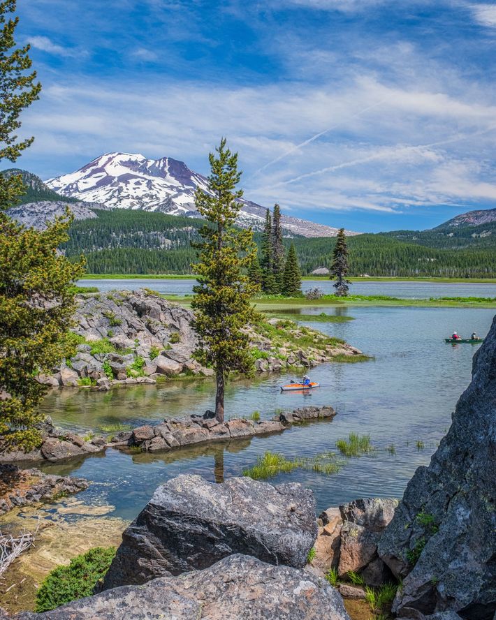 Lake and mountain landscape with a rocky foreground and trees, blue sky with clouds.