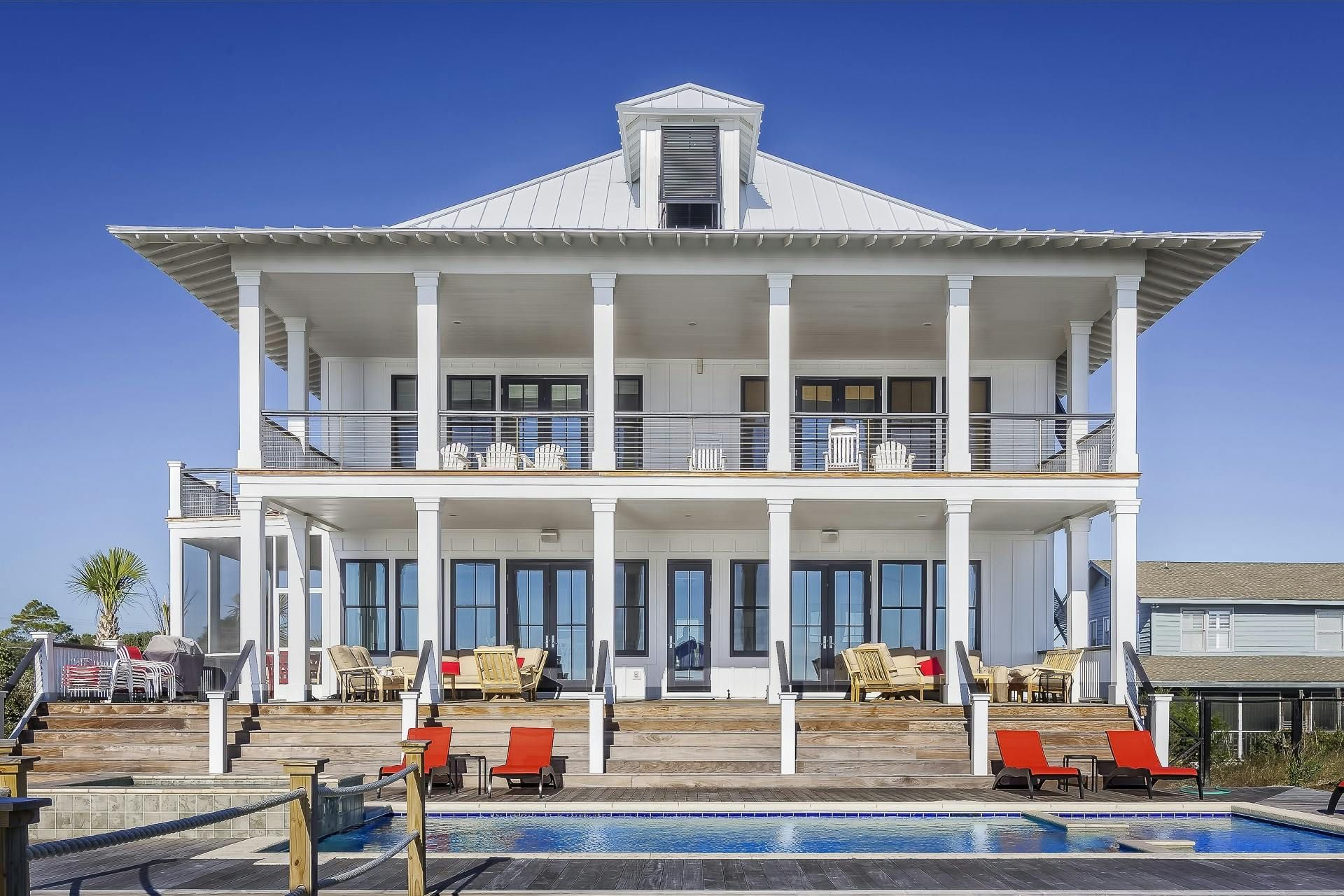 White beach house with multiple levels of balconies overlooking a pool with red chairs. Blue sky in the background.