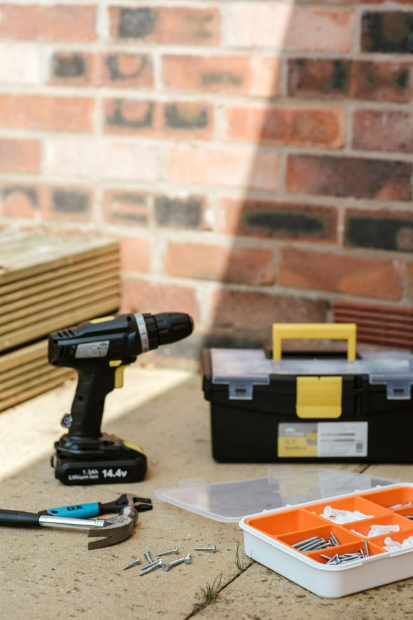 A black drill, toolbox, hammer, and screws on a concrete surface, with stacked wooden planks and a brick wall in the background.