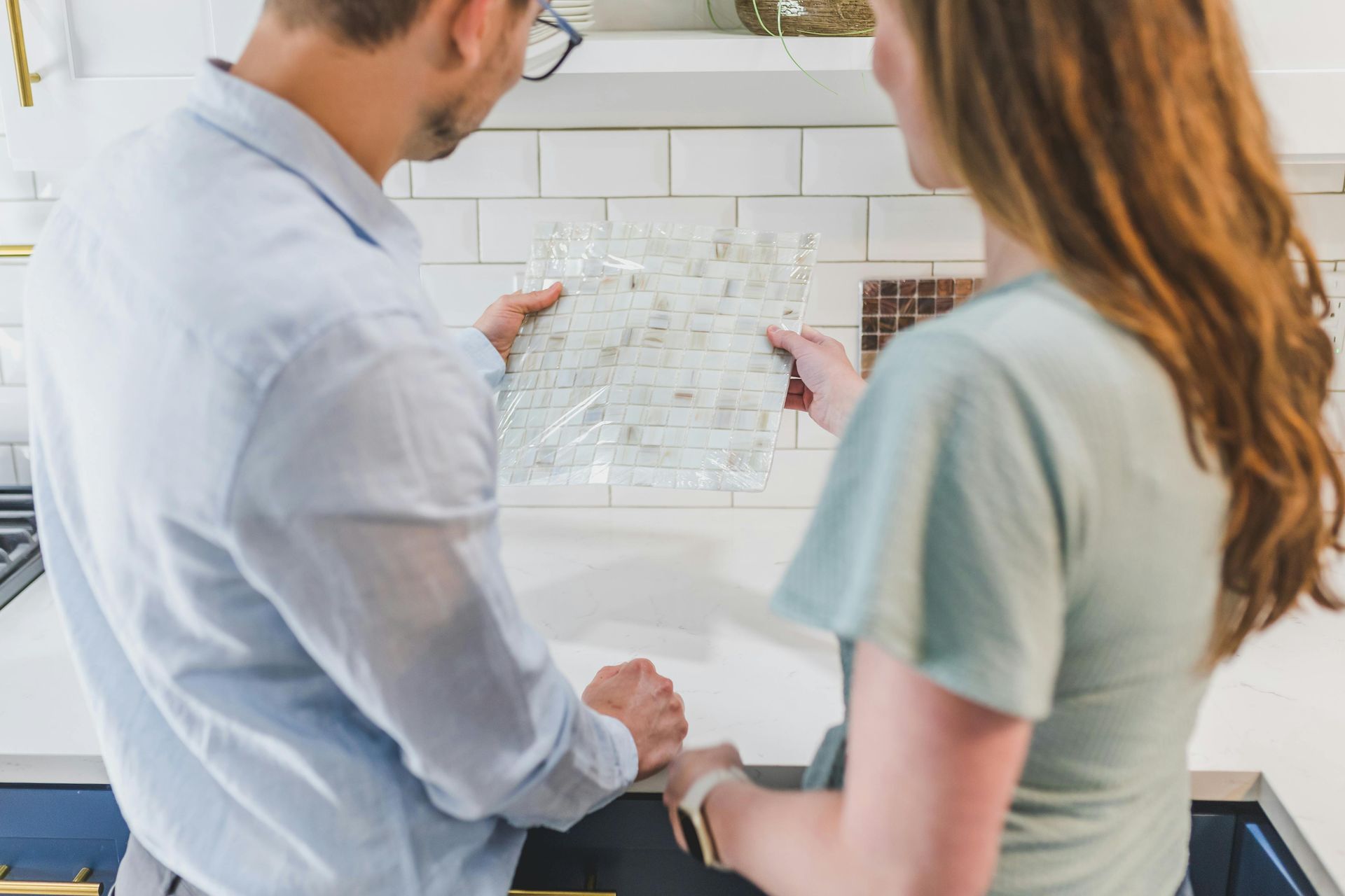 A man and woman in a kitchen examining tile samples. The man wears a light blue shirt, while the woman wears a teal shirt.