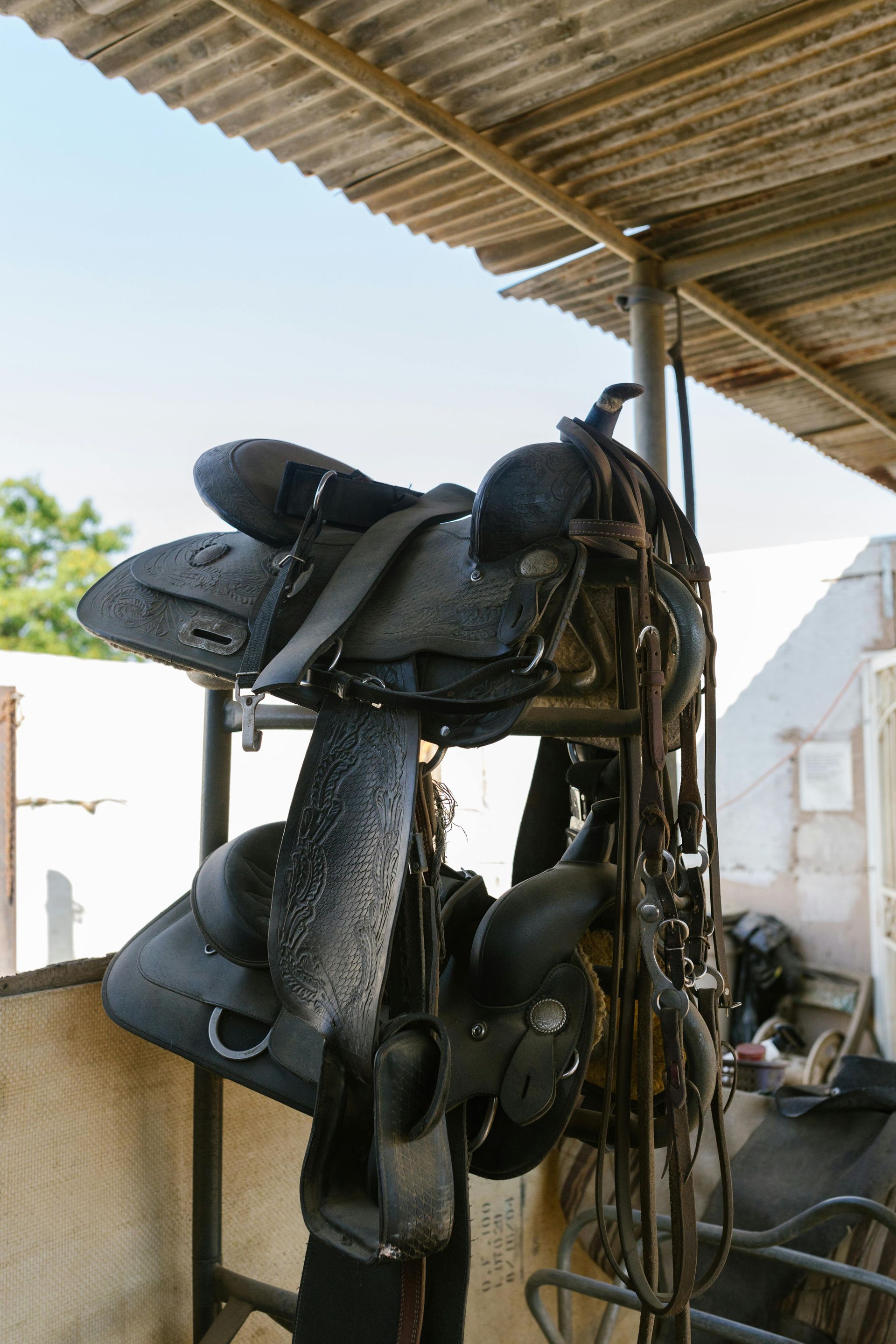 Black horse saddles hanging on a rack under a corrugated metal roof in an outdoor setting.