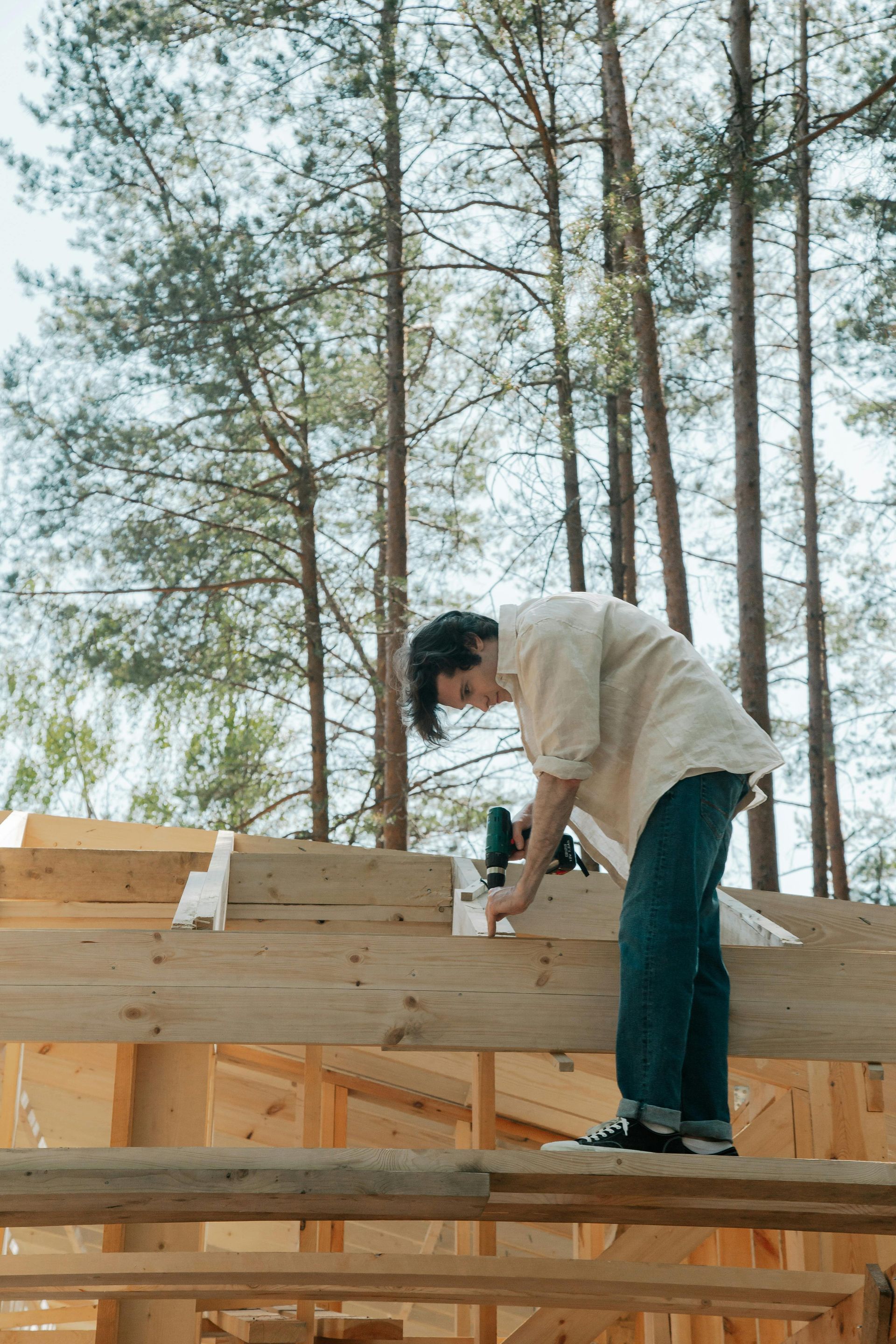 Person in jeans and a cream shirt using a drill on wooden beams, likely building a structure outdoors with trees in the background.